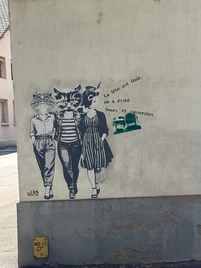 This media file depicts a mural on a light-colored, textured wall, likely an exterior surface. The mural features three figures, rendered in a stencil-like black and white style. Each figure is depicted from the waist down, with their legs and feet visible, and they are wearing clothing that appears to be from a past era, possibly the mid-20th century. The figures are superimposed with animal heads: the top figure has a dog or wolf-like head, the middle figure has a cat head, and the bottom figure has a frog head. The figures are standing close together.

To the left of the figures, in a vertical orientation, is text in French. It reads "La fête est finie, on a évité fours et casseroles." This translates to "The party is over, we avoided ovens and casseroles." There is also a small, green stencil of a figure with a helmet or hat above the text.

In the lower right corner of the image, on a separate grey section of the wall, is a yellow rectangular utility marker. It has text that includes "GAZ," "PE," and alphanumeric codes "MB 272," "041," and "25." This indicates a gas utility marker, likely in France.

The overall scene suggests an urban or semi-urban environment, given the presence of the mural and the utility marker. The lighting implies daytime, possibly with sunlight casting shadows, although the direct light source is not clearly visible. The artistic style and the placement of the mural on a weathered wall suggest it is street art. The presence of the utility marker provides a specific location cue, identifying the area as related to gas infrastructure in Charnay-lès-Mâcon, France.