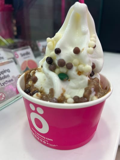 The image shows a close-up of a frozen yogurt dessert in a pink cup. The yogurt is white and topped with a variety of colorful sprinkles, including white, brown, and green. There is a brown chocolate sauce around the bottom of the yogurt. The cup is labeled "ó" with the website "www.emooy.com." The image appears to be taken in a frozen yogurt shop, potentially at the location 43.322, -1.984 in Donostia, Spain.