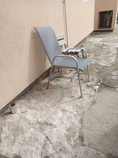An outdoor scene shows two chairs and a metal can on a rough concrete surface next to a building with a beige stucco wall. One chair, a gray metal and mesh lounger, is positioned at an angle to the wall, its legs resting on the concrete. Behind it is a smaller, rustic wooden bench with metal legs. A dented silver metal can is on the concrete to the left of the lounger. The ground is uneven, with patches of dirt and debris. The building has a window with bars in the upper right corner. The scene appears to be in a courtyard or an alleyway.