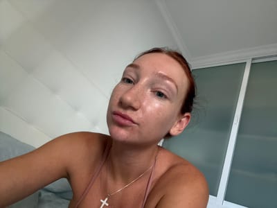 The media file shows a close-up, upside-down selfie of a woman in what appears to be a bathroom or spa-like setting. The woman has fair skin with freckles, blue eyes, and reddish-brown hair. She is wearing a thin, pink strap top and a delicate silver necklace with a cross pendant. Her expression is neutral, and her skin appears slightly dewy, suggesting it might be freshly washed or moisturized.
The background is clean and modern. To the left, there is a white framed window or door with frosted glass panels. To the right and behind her, the surface is a quilted, light-colored material, possibly a spa bed or couch. The overall lighting is bright and even, suggesting an indoor setting during the daytime. There is no visible text in the image. The style of the environment and the woman's appearance suggest a personal care or relaxation context. The image was taken in Las Palmas, Spain.