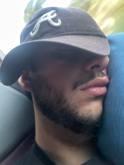 An individual is depicted in a close-up, angled shot, appearing to be asleep or resting. The subject is a male with a dark, trimmed beard and mustache. He wears a dark-colored baseball cap with a light-colored, stylized letter 'A' emblem on the front; the brim of the cap is pulled forward, obscuring his eyes. His head is turned slightly to the left and rests against a blue, soft-textured object, consistent with a cushion or headrest. A dark garment is visible around his neck. The background is out of focus, suggesting an interior environment, possibly a vehicle, with a diffuse bright light source and indistinct green elements visible to the upper right. This scene takes place in Leiria, Portugal, likely during travel or transport within the city or its vicinity.