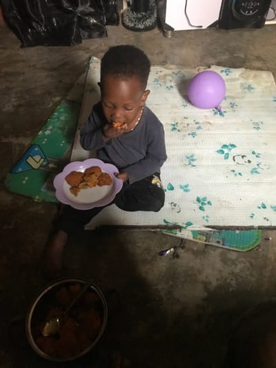 An image taken in Oba-Ile, Nigeria, depicts a young child seated on the floor indoors. The child, a toddler with dark skin and short dark hair, is wearing a dark grey long-sleeved shirt, dark pants, and a light-colored beaded necklace. They are sitting cross-legged on a light-colored foam mat adorned with green floral and panda designs. The child holds a light purple, fluted-edge plate containing several irregularly shaped, fried food items. One piece of food is held to the child's mouth with their right hand, indicating active consumption. Visible around the child's mouth are food crumbs. To the child's right, on the mat, lies a partially deflated purple balloon. In the background, a black plastic bag, a dark speaker-like object, and a white rectangular object are discernible. In the lower left foreground, a metal pot containing more of the fried food items, along with a metal spoon, rests on the concrete floor. The floor is concrete and appears discolored in areas, with another segment of green patterned matting visible to the child's left. Lighting is dim, creating visible shadows.