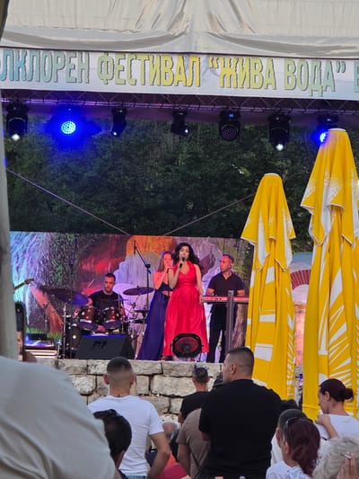 The image captures an outdoor performance at a festival in Gara Oreshets, Bulgaria. A band is on stage, with a female singer in a red dress in the foreground, holding a microphone and singing. To her left, a female in a blue dress stands next to her. A male drummer is seated behind a drum set, and another male is playing a keyboard to the right. The stage backdrop features a colorful, abstract mural. Above the stage, a banner displays text in Cyrillic. Several large, yellow umbrellas with white patterns are visible on the right side of the frame. In the foreground, the backs of an audience are visible, seated and standing, facing the stage. The event appears to be taking place in the evening, with stage lighting illuminating the performers.