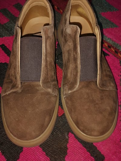A pair of brown suede slip-on shoes are laid out on a vibrantly colored, patterned rug. The rug has sections of bright pink and dark gray, with a geometric design. The shoes are positioned side-by-side, with the toe of each shoe pointing slightly inward. Both shoes appear to be the same size and style, featuring elastic gores on the sides and a rounded toe. The interior of the shoes, visible at the top, is a lighter tan color. The overall scene is a close-up, overhead shot, suggesting the shoes are a focal point. There is no indication of the location being in Juma, Uzbekistan; the image solely depicts the shoes and the rug.