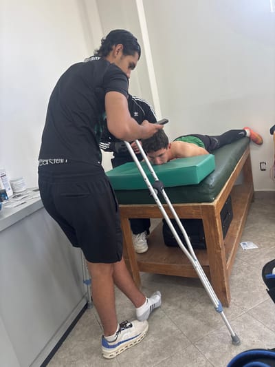 The scene depicts an interior view of a medical examination room or physical therapy clinic located in Las Crucitas, Mexico. The room features white walls and a light-colored tiled floor.
In the left foreground, a male subject with dark hair, wearing a black short-sleeved t-shirt, black athletic shorts, and white athletic shoes with blue accents, stands partially supported by a single silver crutch held under his left armpit. His left foot is grounded, wearing a white athletic shoe, while his right foot is elevated, encased in a white sock with grey patterns. He holds a black smartphone in his right hand and appears to be looking at the screen. The waistband of his Calvin Klein underwear is visible above his shorts. Adjacent to him is a white counter holding various containers and small white packets, suggesting medical supplies.
To the right, a second silver crutch leans against a dark green examination table. On this table, a second male subject, shirtless, with dark hair, lies prone (face down) on a light green rectangular cushion. He is wearing dark athletic pants and orange athletic shoes, with his right leg extended. His face is turned slightly to the right, resting on his forearms. A third individual, wearing a black long-sleeved shirt with white Adidas stripes, is partially visible standing behind the head of the prone subject, suggesting a medical professional or assistant. Their feet, wearing white athletic shoes, are visible beneath the table. Under the table, a black bag rests on a wooden shelf. On the far right wall, a white electrical outlet with visible red and black wires is present.