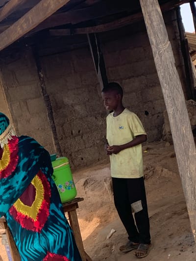 An eye-level, rotated outdoor shot depicts a person standing within a structure. The person is wearing a light yellow t-shirt, black pants, and sandals. They are positioned against a wall. In the foreground, a colorful patterned cloth covers an object that looks like a bed and a bright green plastic container rests on top of the object. The structure appears old and is built with what may be wood and mud, as evidenced by the textures and materials. The location is Tataura, Nigeria.