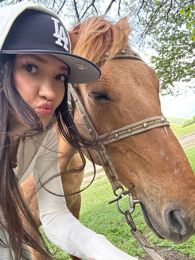 A close-up, angled selfie depicts a light-skinned woman wearing a dark blue baseball cap with the "LA" logo in white, a white hooded sweatshirt, and dark hair. The woman is making a kissing face toward the camera. Adjacent to the woman is a light brown horse with a decorative bridle. The background includes green foliage and what appears to be an open field. It is difficult to determine the specific location within the city of Teghut, Armenia from the given image.