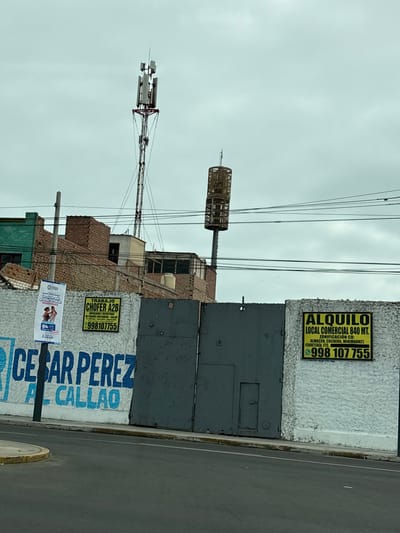This is a vertical, medium-shot photograph of a street scene in Callao, Peru. The sky is overcast and a pale grey, suggesting a cloudy or possibly rainy day. The foreground features a street and a sidewalk.
Dominating the right side of the image is a rough, textured white wall. On this wall, there are two prominent signs. The upper sign, in a bright yellow rectangle with black lettering, advertises "ALQUILO LOCAL COMERCIAL 840 MT." (RENT COMMERCIAL SPACE 840 SQM). It lists potential uses such as "ALMACÉN, COCHERA, MINIMARKET, FERRETERÍA, ETC." (WAREHOUSE, GARAGE, MINIMARKET, HARDWARE STORE, ETC.) and provides a phone number: 998 107 755. Below this, a large blue stencil reads "CESAR PEREZ CALLAO," likely a political or campaign message.
To the left of the wall, various structures are visible, including brick buildings in disrepair and several tall communication towers or antennae. One tower has a cage-like structure near its top. Another sign on a white pole below the main wall advertises "TRABAJO CHOFER A2B" (JOB DRIVER A2B) and "EMPACADOR Y REPARTIDOR BE PEDIDOS PORTUARIOS-CALLAO" (PACKER AND DELIVERY PERSON FOR PORT ORDERS-CALLAO), also with a phone number: 998107755. A smaller banner below that features an image of two people dancing salsa and the text "La clase más grande de salsa CALLAO" (The biggest salsa class CALLAO). Overhead, a network of electrical wires crisscrosses the sky. The overall impression is one of urban infrastructure, commercial activity, and local communication in a cloudy environment.