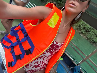 A woman, possibly on a boat in Đồng Văn Commune, Vietnam, is wearing a life vest. The vest is bright orange with blue straps and black buckles. She wears a white bathing suit with a red pattern, long silver earrings, and sunglasses. The boat's interior has green and white-striped surfaces, and the surrounding environment includes green foliage and water. Another person's legs and shoes can be seen partially in the bottom right corner.