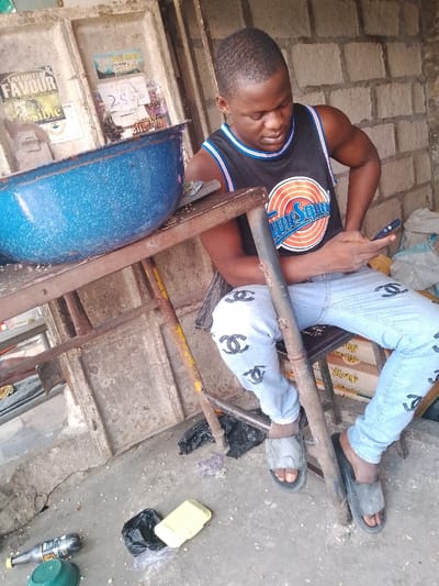 The image portrays a young man seated in what appears to be an informal, outdoor or semi-enclosed setting, typical of everyday life in Mgbuoshimini, Nigeria. He is positioned next to a rusty metal table that holds a large, blue speckled enamel basin containing granular material. The background consists of a rough concrete or brick wall adorned with several posters, one prominently displaying "UNLIMITED FAVOUR" and another partially visible with the word "FAITH".

The man has short, dark hair and is wearing a black sleeveless tank top featuring an orange and white circular logo with "Tune Squad" written on it. He is dressed in light blue jeans embellished with multiple black Chanel logos, and is wearing grey slide sandals. His gaze is directed downwards, focused intently on a small, dark-colored mobile device held in his hands, indicating he is actively engaged with it. He appears calm and focused.

The ground, a concrete surface, is scattered with various items of debris, including a black plastic bag, a yellow foam container, a plastic bottle, and a green plastic bowl. The bright lighting suggests it is daytime. The scene captures a candid moment of the man engrossed in his device within a functional, unadorned environment.