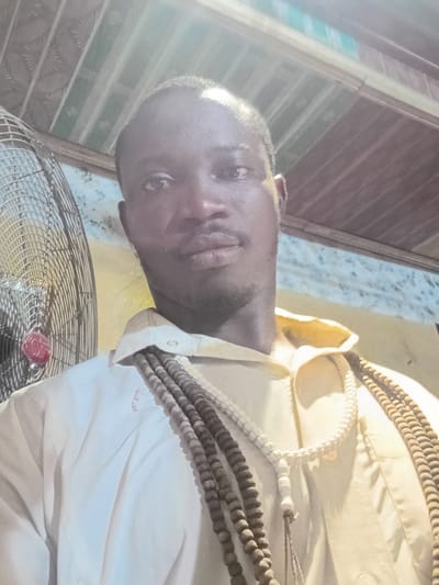 A selfie taken from a slightly upward angle shows a Black man from the chest up. He has dark skin and is wearing a light-colored collared shirt. Around his neck, he wears several strands of prayer beads, varying in size and color from light beige to dark brown. To the left of the man, a large, metallic fan with a red accent is visible. The background suggests an indoor setting, with a corrugated metal ceiling above and a patterned wallpaper or wall covering behind. The lighting is bright, casting some glare and shadows.