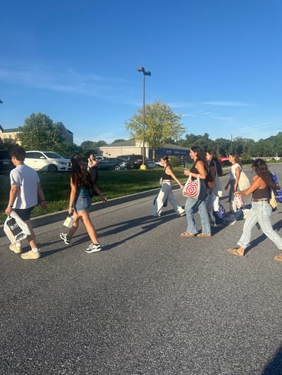 A group of young people, predominantly female, are crossing a paved road in a crosswalk. The scene appears to be in an outdoor commercial or suburban area, possibly a parking lot or access road. The sky is clear and blue with some wispy clouds, and the sun is casting long shadows, indicating it is late afternoon or early evening.

In the foreground, from left to right, a male individual wearing a white t-shirt and shorts is walking with a white shopping bag. Next to him, a female in a teal top and denim shorts is also walking, carrying a small white bag. Further ahead, a group of women are walking together, some carrying shopping bags, including one with a red and white circular logo that resembles a Target bag. Another woman in the group is holding a smartphone. The people are dressed in casual summer attire.

In the background, there are buildings that resemble commercial establishments or a hotel, and several parked cars, including a white sedan and a dark-colored sedan. Trees and some shrubbery line the area behind the road. A street lamp is visible in the background. The overall impression is of a casual outing, possibly involving shopping, on a pleasant day.