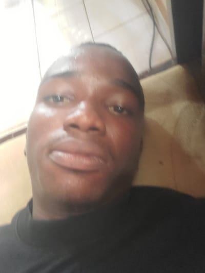 The image is a close-up selfie of a Black male wearing a black t-shirt. He is lying down, with his head tilted back and facing the camera. The background appears to be indoors, with tiled walls and flooring visible. The lighting is bright and somewhat washed out, creating a glare on his face and making some details difficult to discern. The man's facial features are prominent, with his eyes open and looking towards the camera, and his lips slightly parted. The overall impression is of a casual, personal snapshot taken in a domestic setting. There are no obvious indications of the specific location within Oturkpo, Nigeria, beyond it being an indoor environment.