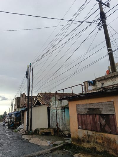 The image depicts a street scene in Sindangjaya, Indonesia, under an overcast grey sky. Multiple utility poles are visible, primarily along the left side of the street, supporting a dense and tangled network of black electrical and communication wires that crisscross the frame. One pole on the far right features a substantial cluster of bundled wires and equipment.
The street itself is dark, likely asphalt, appearing uneven and possibly damp, with small puddles and patches of lighter ground near the building foundations.
Buildings line the street:
* On the far left, a white van and two indistinct human figures are visible down the road; one appears standing, the other near a cart or bicycle. Adjacent to the road is a small street stall with a blue tarp roof.
* Further along the left, several multi-story structures are visible, some appearing incomplete with exposed concrete frameworks and unrendered walls. A building with a dark, tiled gable roof is situated in the mid-ground.
* Moving right, a single-story building with a light beige facade is present, next to a weathered structure with a light blue-grey surface. A concrete bench-like structure is located in front of the beige building.
* In the foreground on the right, a single-story building features a faded orange/ochre facade. Its window opening is boarded with rough, horizontal dark wooden planks. The lower portion of this building's wall exhibits dark green/black staining or biological growth. On the roof of a building immediately behind this structure, a red water tank, metal railings, and clothes hanging on a line are visible.