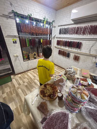 The scene depicts the interior of a small retail establishment in Manavi, Georgia, specializing in traditional Georgian sweets and snacks. A young child, viewed from the back, stands centrally, wearing a yellow t-shirt with "football school 35" printed in black on the rear, observing the products.
The walls are covered with white brick-patterned material, and the ceiling is paneled with light-colored wood. A large window on the left side is adorned with string lights and artificial grapevines, with bottles resting on the windowsill. Below the window, a white electrical outlet has a white and black cable plugged in, and a yellow QR code sticker is affixed to the wall.
Numerous strings of Churchkhela, a Georgian nut candy, hang from horizontal rods across the window and along the adjacent right wall. These display various colors, including brown, dark red, green, yellow, and black. Above these displays on the right wall, a white split-unit air conditioner is mounted.
A long table, covered with a white cloth, occupies the foreground. On the table, a wooden platter holds an assortment of walnuts and dried fruits. A large transparent plastic bag filled with walnuts is centrally placed. Several items of Tklapi (fruit leather), some circular and others rectangular, are wrapped in plastic. A stack of colorful, woven baskets contains small, individually packaged items, possibly spices or small sweets. Further right on the table, a stack of flat brown paper bags, pink scissors, a roll of packaging material, and a black calculator are visible. A white-handled knife rests on the table.
On the far left, a partial view of a doorway shows a dark-colored interior beyond and a green mat on the light-colored, patterned floor. The floor itself consists of light brown and beige rectangular tiles arranged in a staggered pattern.