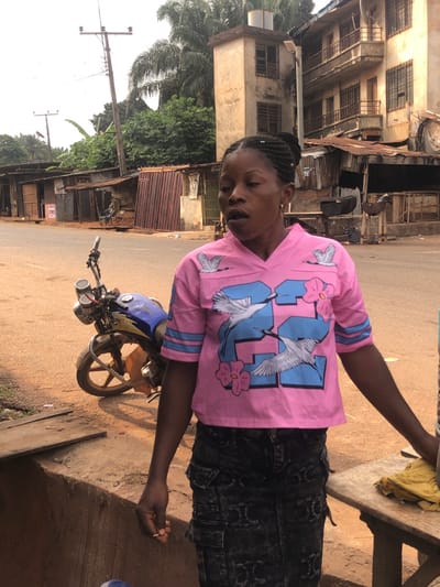 This image depicts a street scene in Neni, Nigeria. A dark-skinned female, with braided hair pulled into an updo, stands in the foreground, slightly right of center. She wears a pink short-sleeved V-neck shirt adorned with blue stripes on the sleeves, a large blue "32" graphic, and white crane and pink flower designs. She is also wearing a dark, distressed denim cargo skirt. Her mouth is slightly open, and her gaze is directed downwards and to the right of the frame. Her left hand loosely holds a small, light-colored, cylindrical object. To her immediate right, a portion of a wooden surface, possibly a table or counter, is visible.
In the mid-ground to the left, a blue and black motorcycle is parked on a reddish-brown dirt shoulder adjacent to a paved road. The background features a blend of structures and vegetation. On the left, a series of single-story buildings, characterized by corrugated metal roofs and varied wall materials, line the street. On the right, a multi-story concrete building, appearing weathered, stands prominently, alongside a taller, narrower concrete structure. Dense green trees and palm foliage are visible behind these buildings. Several utility poles with overhead wires are present against the bright sky. The immediate foreground consists of reddish-brown dirt, with a low concrete barrier or drainage ditch on the left side.