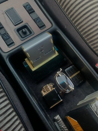 Within a vehicle, purportedly located in Chonas-l'Amballan, France, the image depicts a close-up, high-angle view of a dark interior storage compartment and an adjacent control panel. The storage compartment, lined with dark felt, contains several items. A silver and gold-trimmed perpetual calendar unit displaying "4," a starburst symbol, "AVRIL," and "MERCREDI" rests on a dark, reflective base in the upper-left of the compartment. To its right are a black and gold Zippo-style lighter and a rectangular black lighter with a textured pattern and gold accents. Between these lighters, a pair of silver-framed, folded reading glasses is positioned. In the lower-left of the compartment, a silver, ridged utility knife or multi-tool is partially visible, alongside a small, partially obscured orange tag. The adjacent control panel features a faux wood-grain finish, housing four black rectangular power window switches (two on the left, two on the right) and a central black toggle switch within a recessed square. Portions of striped fabric upholstery, with light and dark bands, are visible along the right and bottom edges of the frame. No individuals are present.