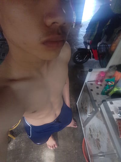A shirtless person with blue shorts is standing on a concrete floor. To the right, there is a white cabinet with drawers. On top of the cabinet, there are several items including a pink water bottle and a green packet. Behind the person, there is a black motorcycle with red accents. Clothing is hanging on a rack in the background. The scene appears to be indoors, possibly in a residential setting in Pompanua, Indonesia, judging by the available context.