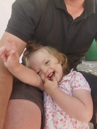 A child, with light brown hair and fair skin, is held on a person's lap. The child is wearing a light pink dress with a floral pattern. The child is smiling broadly, with their fingers near their mouth. The adult, wearing a dark polo shirt and dark shorts, is partially visible in the frame. The background suggests an outdoor setting, possibly a patio with patterned sunlight on the ground. The image appears to have been taken in Carbuna, Moldova, as indicated by the context.