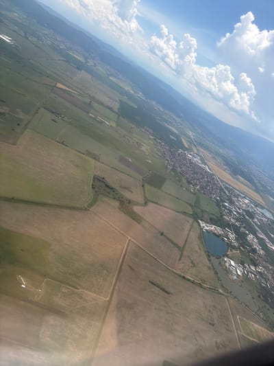 An aerial view captures a landscape dominated by agricultural fields and a town, likely Dolni Bogrov, Bulgaria. The scene is tilted, suggesting it was taken from an airplane. A prominent feature is a series of cultivated fields, appearing in varying shades of green and brown, divided by thin dirt paths or roads. In the middle ground, a town is visible, characterized by its densely packed red-tiled roofs and a main road running through it. Adjacent to the town, a body of water, possibly a lake or reservoir, is present, with some buildings situated along its edges. Further into the distance, mountains form a backdrop under a bright blue sky filled with fluffy white clouds. The overall impression is one of a rural-urban interface, where agricultural land meets a developed settlement against a natural mountainous setting.
