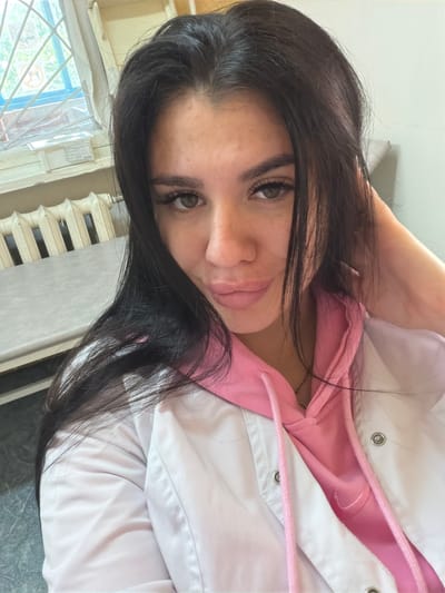 A young woman with long, dark hair is taking a selfie in what appears to be a room with a window and a radiator. She is wearing a pink hooded sweatshirt under a white jacket. The window has blue panes and a white frame, and through it, greenery can be seen. A white radiator is positioned below the window. A table with a textured surface is visible to the left of the woman. The overall lighting suggests an indoor setting. There is no specific visual information in the image to indicate the location within the city of Чумна, Russia.