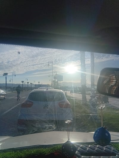 A view from inside a vehicle captures a street scene in Martil, Morocco. A Kia car is visible through the windshield, parked alongside a road lined with palm trees and decorative street lamps. A person is walking on the sidewalk. The sun is shining brightly in the distance, creating a glare. The scene includes elements of urban infrastructure and a pedestrian walking along the street.