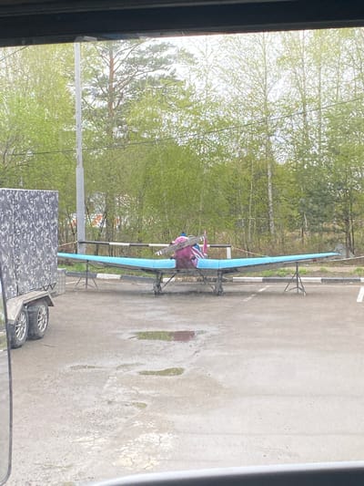 The image depicts a hang glider atop a trailer, parked on an asphalt surface. The surroundings consist of a treeline and a utility pole. The location is identified as Молодежный, Russia.