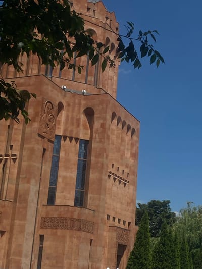 The image captures a detailed view of the Surp Astvatsatsin Church (Holy Mother of God Church) in Nor Hachin, Armenia, under a clear blue sky. The prominent structure is a multi-story church built from reddish-brown stone, likely tuff. A large, dark green leafy tree partially obstructs the upper left and central portions of the building, with its branches extending into the upper right.

The church features a tall, cylindrical upper section with multiple arched openings, indicating a dome or tower. Below this, the main body of the building displays tall, narrow windows with dark glass, some set within arched recesses. A notable architectural detail is a bas-relief carving on an upper wall section, depicting two figures, identifiable as the Virgin Mary holding the Child Jesus. Further down, an intricate carved frieze with floral or geometric patterns runs horizontally below a series of windows. Small, triangular projections are also visible as decorative elements on some wall surfaces.

In the lower right corner, several tall, dark green conical evergreen trees are visible, with a backdrop of lighter green deciduous trees further behind them. The scene is brightly lit by natural daylight, casting subtle shadows on the building's facade. No people are present in the image.