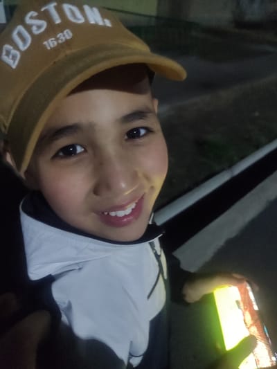 A close-up, angled portrait captures a young individual, likely male, smiling directly at the camera in Rishtan, Uzbekistan. The subject wears a light brown baseball cap with "BOSTON." and "1630" embroidered in white text on the front, along with a white and dark-colored (possibly black or dark blue) jacket. The left hand (viewer's perspective) holds a smartphone with an illuminated screen displaying digital content, emitting a yellow-green glow onto the hand and lower face. The background is dark, suggesting nighttime or low light conditions, with a visible curb or raised edge in the lower right portion of the frame.