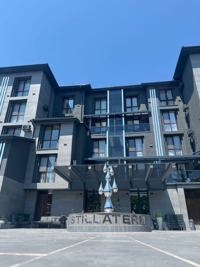The image shows the exterior of a modern hotel or resort building in Mineralni Bani, Bulgaria. The building has a dark gray facade with several stories of windows and balconies. A contemporary sculpture, appearing to be made of polished chrome droplets, is positioned in front of the entrance. The name "STILLATERM" is visible on a curved structure at the base of the sculpture. In the foreground, there is a paved outdoor area with some seating. The sky is clear and blue.