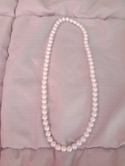 A single strand of white faux pearls is laid out in a curved shape on a soft, light purple quilted surface. The pearls are uniform in size and shape, strung together on thin white thread. The lighting is soft, casting subtle shadows. The scene appears to be indoors. There are no people or visible actions. The location in Inhulo-Kamianka, Ukraine, is not discernible from the image.