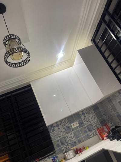 This media file captures an indoor scene, likely a kitchen or utility area, within a residence in Adenta Municipality, Ghana. The image is taken at an angle, resulting in a tilted perspective.
The room features clean white walls adorned with decorative crown molding and a white ceiling. A section of a black grid structure, possibly a decorative ceiling element or part of a suspended ceiling, is visible in the upper left. The space is illuminated by two light sources: a circular recessed light fixture emitting a bright glow on the wall, and a decorative hanging pendant light in the lower left, featuring a black metal cage surrounding a cylindrical glass shade.
A significant portion of the image is occupied by modern, glossy white L-shaped cabinetry, likely kitchen cupboards, which reflect the light. The floor or a backsplash area is covered with distinct patterned tiles in a mix of light blue, grey, and white, showcasing various geometric and abstract designs. A white electrical outlet with a switch is mounted on this tiled surface.
On the tiled surface, towards the upper right, a black plastic bag, a red and white packaged item (partially showing "KIVO" text, suggesting a brand name), and other small items are visible. Further down, a stainless steel pot or bowl and more small packaged goods are present. In the lower right, a dark, black metal-barred structure, possibly a window with security bars or a door, is seen. The prominent artificial lighting suggests the time of day could be evening or night, or that the interior requires constant illumination. No individuals are present, and no specific activity is taking place.