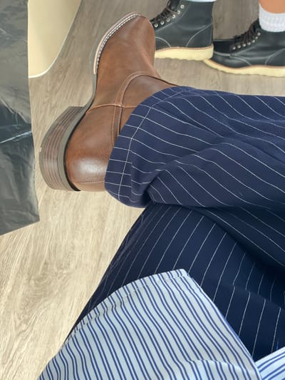 A close-up, overhead shot shows a person's legs crossed, resting on a light-colored wooden floor. The person is wearing dark navy pants with thin white pinstripes and a brown leather boot. In the background, another pair of black boots with light tan soles and white stitching is visible, along with white socks. The environment appears to be indoors, possibly a room with wooden flooring. No specific landmarks or features indicating the city of Simagino, Russia, are present in the image.