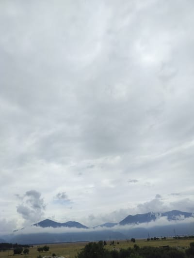 This is a landscape photograph taken in Gorno Sahrane, Bulgaria. The image depicts a cloudy sky that fills the majority of the frame. Below the sky, a range of mountains can be seen, partially shrouded in mist and clouds, giving them a soft, diffused appearance. The mountains are dark blue, indicating distance or the overcast weather.
In the foreground, there is a vast, open field with dry, yellow grass, suggesting it might be late summer or early autumn. Scattered trees are visible in the middle ground, along with some power lines and pylons, hinting at human presence and infrastructure in the rural setting. The overall mood of the image is somber and tranquil, dominated by the overcast sky and the muted colors of the landscape. The time of day appears to be daytime, but the heavy cloud cover obscures direct sunlight, leading to diffused lighting conditions. There is no visible text in the image.