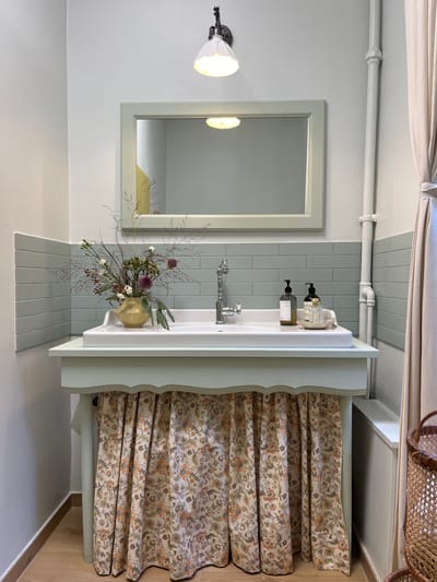 The image displays an interior washroom or powder room, situated in Kaiserslautern, Germany. A white rectangular ceramic sink is integrated into a light green painted wooden vanity. The vanity's front features a scalloped trim and is draped with a pleated fabric skirt, patterned with orange and green floral motifs on a light background.
A chrome, traditionally styled single-handle faucet is installed centrally at the back of the sink. To the left of the faucet, a golden-yellow pitcher-shaped vase holds a rustic floral arrangement containing dried grasses, small red berries, white blossoms, and prominent dark purple spherical flowers. To the right of the faucet, two dark-colored pump bottles, likely soap and lotion dispensers, rest on a small light-colored tray, alongside a small white ceramic cup.
Above the sink, a rectangular mirror with a light green wooden frame is mounted. A wall-mounted light fixture with a white fluted glass shade and a dark metal arm is positioned directly above the mirror, with its bulb illuminated. The wall area behind the sink and mirror is covered with a backsplash of horizontal light teal subway tiles. The upper wall sections are painted a light, neutral color.
On the right side of the scene, a light beige curtain is tied back, revealing a white painted vertical pipe with several horizontal sections running down the wall. A partial view of a dark brown wicker basket is visible in the bottom right corner. The flooring consists of light-colored wooden planks. The mirror's reflection indicates an interior space with a visible section of a yellow wall.