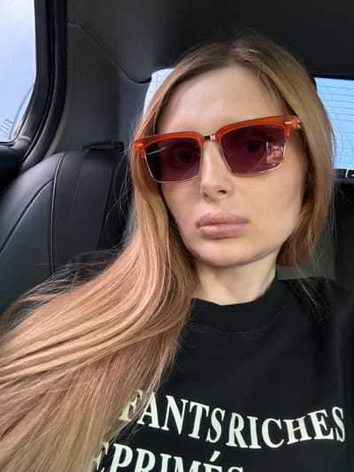 The image is a close-up, portrait-style photograph of a young woman taken from a high angle. She is wearing oversized, translucent red-tinted sunglasses with thick, angular orange frames. Her fair skin is visible, and her lips are slightly parted. Her long, blonde hair, with hints of strawberry blonde, is fanned out across the frame, partially obscuring her left shoulder and the black crewneck sweatshirt she is wearing. White text is printed vertically on the sweatshirt, with visible words including "NTS RICHES" and "PRIME." The woman appears to be sitting in the passenger seat of a car, as indicated by the dark, textured upholstery of the car's interior visible in the foreground and the side of the car's headrest. The background is slightly blurred, showing the car's ceiling and a sliver of the sky through a window, suggesting it is daytime. The lighting is somewhat dim, possibly indicating an overcast day or a shaded interior. The woman's expression is neutral, and her gaze is directed slightly away from the camera. The overall scene suggests a casual moment, possibly a car ride. Visible text on the car window includes "ATOTOT." The location context is Starobzhegokay, Russia.