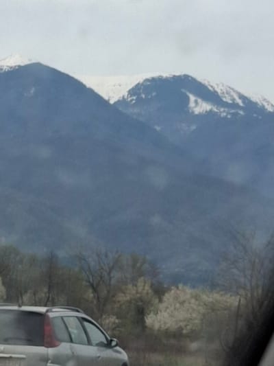The image shows a mountain with snow on its peak next to a road with trees and a car. The image was taken near Mezdreya, Bulgaria.