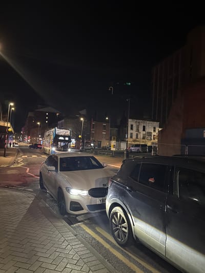 The image captures a nighttime urban street scene in Preston, United Kingdom. In the foreground, a white BMW sedan with a visible driver is positioned in extremely close proximity to the rear bumper of a dark grey SUV (likely a Volkswagen T-Cross) on its right. The BMW's front passenger wheel is on the block-paved sidewalk, while the rest of the vehicle extends into the road near double yellow lines. The SUV is on the road, adjacent to the curb, and displays a red and white hanging ornament in its rear side window. Both vehicles have their headlights activated.
In the mid-ground, a Stagecoach route 125 double-decker bus is visible on the road, approaching from the background. Surrounding the street are various urban buildings under artificial light: commercial establishments on the left, a dilapidated multi-story building with dark window openings on the right, and a large, imposing, dark building with vertical architectural elements on the far right. Streetlights illuminate the scene, and traffic signals are discernible. The sky is dark.