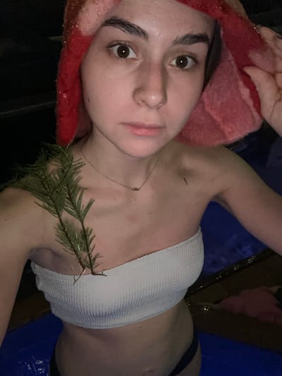 A female individual, appearing to be in her late teens or early twenties, is depicted in a medium close-up, partially submerged in water, presumably a pool or hot tub, at night or in a dimly lit indoor setting in Базарово, Russia. She is wearing a white, ribbed strapless bikini top and dark-colored bikini bottoms. Her head is wrapped in a textured, bright red fabric head covering, possibly sparkling or sequined. She gazes directly into the camera with wide-open eyes and a neutral expression. A thin, silver-colored necklace with a small, dark pendant is visible around her neck. Two green coniferous branches are positioned on her left shoulder and upper chest, tucked into the top of her bikini. A small, dark, linear object is visible on her right clavicle area. The water surface is blue, and the background is dark and indistinct.