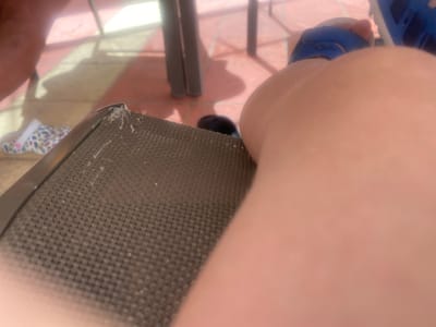 The image shows a close-up view of a person's leg resting on a chair. The person is wearing a blue sandal. In the background, there's a chair with a woven seat and metal legs. The floor is tiled in a terracotta color. There is a patterned shoe visible on the floor in the periphery. The location is speculated to be in Monastir, Tunisia.