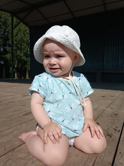 A young child, wearing a white bucket hat and a light blue t-shirt with dragonfly and rainbow patterns, sits on a weathered wooden deck. The child is facing to the left with a slight grimace, showing a bit of discomfort. Their hands are resting on their thighs. In the background, trees are visible, and a dark, corrugated metal structure with a dark roof stands behind the child. The scene appears to be outdoors, possibly in a park or a recreational area, on a sunny day. The location is in the city of Posad, Russia.