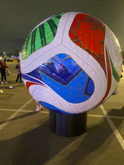 The image shows a giant, inflatable soccer ball on display outdoors. The ball is decorated with white panels interspersed with red, blue, and green sections featuring star and leaf patterns. It is supported by a dark cylindrical base.
Several people are visible around the ball, appearing to admire or interact with it. Some are standing, others are walking. The ground is asphalt, marked with white lines, suggesting a parking lot or an open public space. The time of day appears to be evening or night, as indicated by the artificial lighting and shadows. The weather is not clearly discernible but seems calm.
In the background, there are lights and some structures that suggest a commercial or entertainment venue, possibly a stadium or fairground. The overall mood seems festive and casual. No visible text is present on the soccer ball itself, but some distant signage might be present in the background. The scene takes place in Inglewood, USA.