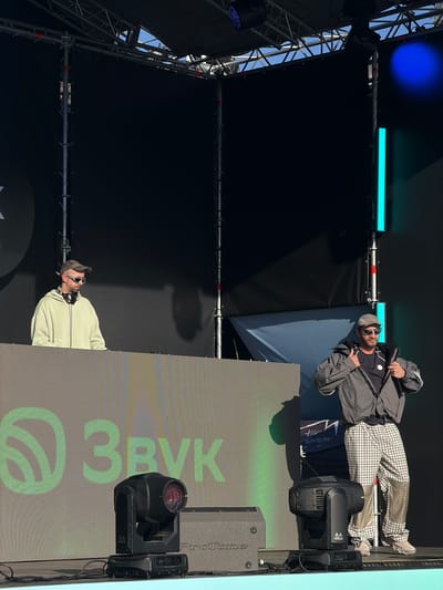The image shows two men performing on a stage. The location is likely Озёрное, Russia, given the "Звук" logo on the screen, which is a Russian word meaning "sound." One man is wearing a light-colored jacket and headphones, while the other is wearing a grey jacket and checkered pants. They are both wearing sunglasses and holding microphones. The stage has lighting equipment and a backdrop with the "Звук" logo.