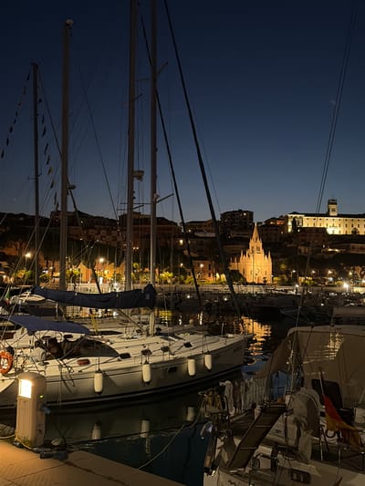 At dusk in the city of Imperia, Italy, a harbor is illuminated by artificial lights. Numerous sailboats are moored in the foreground, their masts and rigging creating a complex silhouette against the darkening sky. The hulls of several boats are visible, with buoys attached to their sides. One boat in the immediate foreground has a blue cover over its boom. A power pedestal with a light is situated on the dock next to this boat. In the background, a city skyline rises, featuring buildings and a prominent church with a spired roof, all softly lit. The reflections of the lights shimmer on the water's surface. The sky is a deep, clear blue, transitioning to dark.