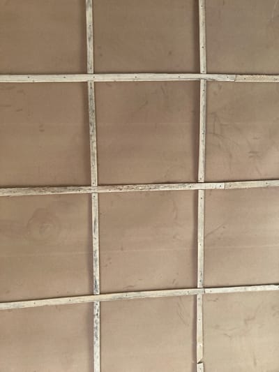 The image shows a close-up of the back of a large framed surface. The frame is constructed of narrow, light-colored wooden strips arranged in a grid pattern. The grid divides a brown surface into twelve rectangular sections. The wooden strips are attached to the brown surface with small nails, visible along the wood. The brown surface appears to be a type of board or panel. The image does not contain any information that connects it to Yan Dala, Nigeria. The scene does not include people, actions or interactions.