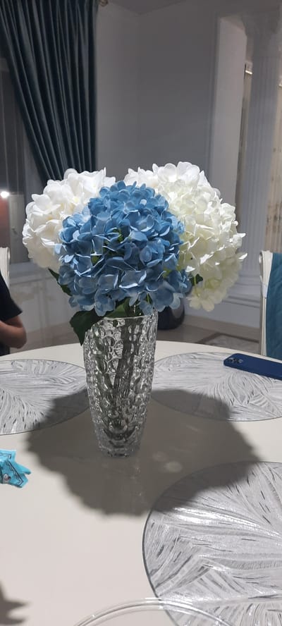 A close-up, vertical shot focuses on a glass vase filled with artificial hydrangeas. The vase, textured with a bubble-like pattern, is positioned on a round, light-colored table. The hydrangeas are arranged in a cluster, with blue blooms at the forefront and white blooms forming the outer layers. Two ornate, circular placemats are visible on the table, one beneath the vase and another to the right. A blue smartphone rests on the placemat to the right. In the background, dark teal curtains hang on the left side of the frame. To the right of the curtains, a white column is visible, and beyond that, a hint of patterned drapery. A person's arm and shoulder, clad in a dark shirt, are visible on the far left, partially obscuring the scene. The lighting suggests an indoor setting, possibly during the evening. There is no visible indication or context suggesting this scene is located in Gudermes, Russia.