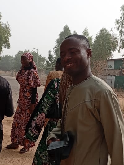 A man smiles at the camera, wearing a light brown, long-sleeved shirt with a decorative trim. He holds a small, black leather bag. Several people are walking nearby, including a woman in a patterned long dress and head covering. Behind them are trees, a street, and a building with a corrugated metal fence. The sky is overcast, giving the scene a muted light.