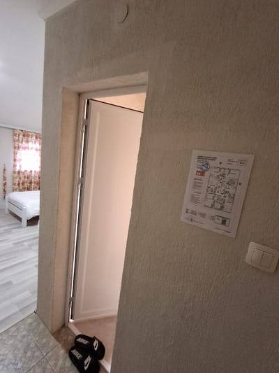 The image depicts a room in Krumovgrad, Bulgaria. The view is looking upwards towards the ceiling, which is textured and off-white in color. A light switch is visible near the top of the frame, along with a printed evacuation plan attached to the ceiling. A rectangular opening in the ceiling leads to another room. Through this opening, a white bed and a window with patterned curtains can be seen. A pair of black Nike-branded slides is on the floor near the opening. The tiles on the floor are light gray.