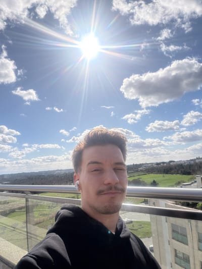 An outdoor portrait, likely a selfie, is taken from a high vantage point, characteristic of a balcony or terrace, in Viseu, Portugal. The primary subject is a male in his 20s or 30s, positioned in the lower half of the frame, looking directly at the camera. He has light brown hair, a mustache, and a short beard. He wears a black hooded sweatshirt and a single white wireless earbud in his left ear. His left eye is partially closed, possibly squinting from the bright overhead sun, which is intensely visible in the upper left quadrant of the image, producing a prominent starburst lens flare against a clear blue sky with scattered white cumulus clouds. Behind the subject, a modern glass and metal railing extends horizontally. The background reveals a panoramic view of Viseu, encompassing rolling green hills, dense tree cover, cultivated land, and several multi-story buildings with light-colored facades.