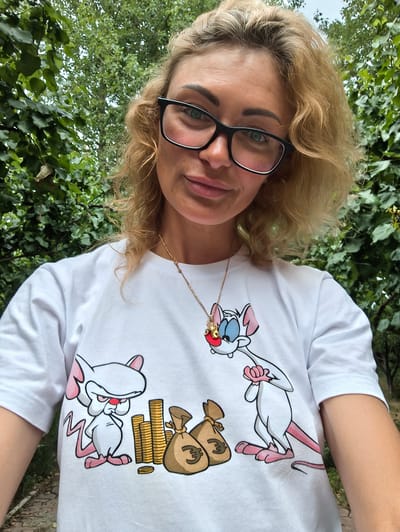 A woman with blonde, curly hair is taking a selfie outdoors. She is wearing black-rimmed glasses and a white t-shirt with a cartoon design of two pink mice, stacks of gold coins, and money bags. One mouse is looking thoughtfully at the money, while the other is standing with its arms crossed. The woman has a neutral expression. Behind her, lush green foliage is visible, suggesting a park or garden setting. The lighting indicates it's daytime, possibly overcast, as there are no harsh shadows. The woman is wearing a gold necklace with a small charm.